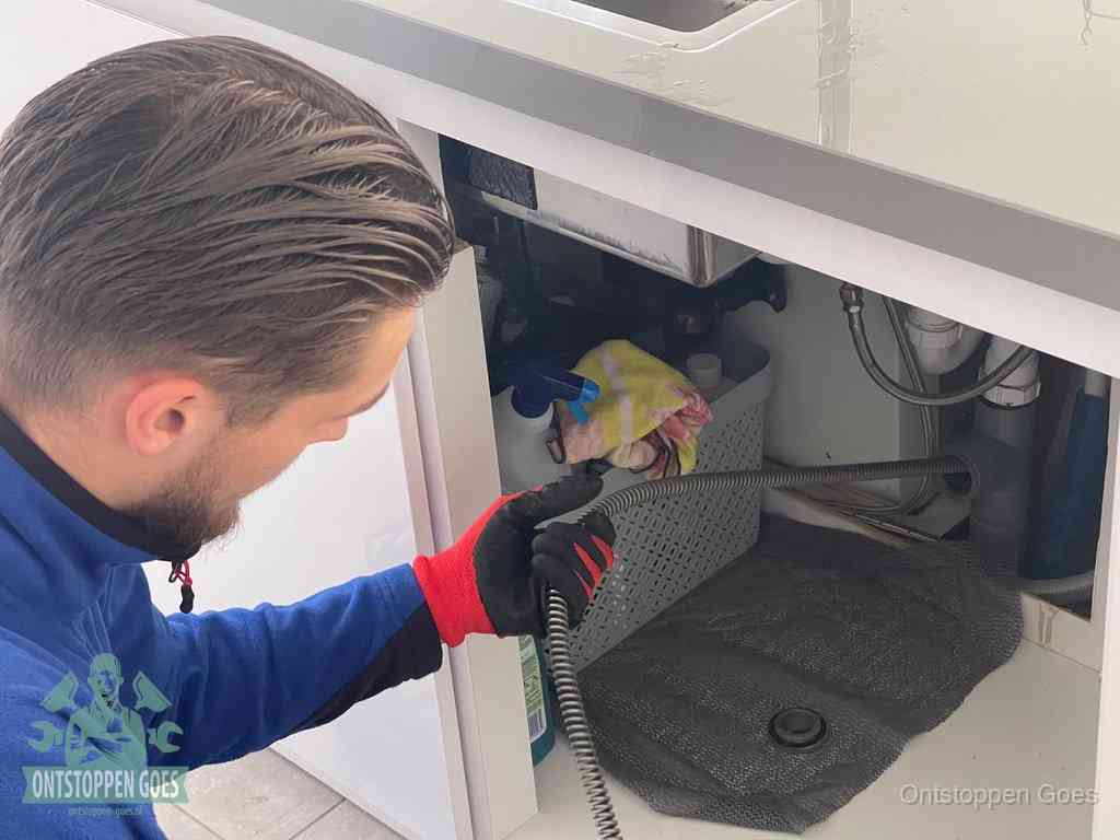 Een monteur voert een flexibele ontstoppingsveer in onder een keukenkastje om een verstopping voorbij de sifon te bereiken in Goes