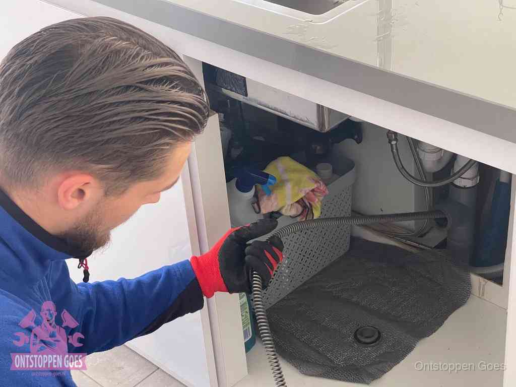 Een monteur voert een flexibele ontstoppingsveer in onder het keukenkastje, langs de sifon, om de gootsteen te ontstoppen in Goes
