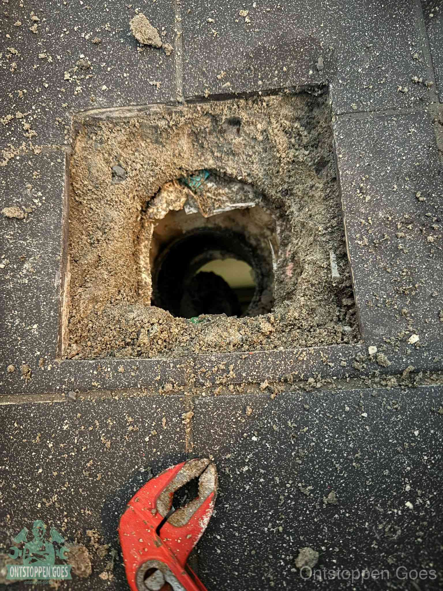 Een geopend rioolputje in een betontegelvloer met losgemaakt rooster en een zichtbare ronde afvoerbuis, het achterhalen waar de afvoerverstopping zit in Goes