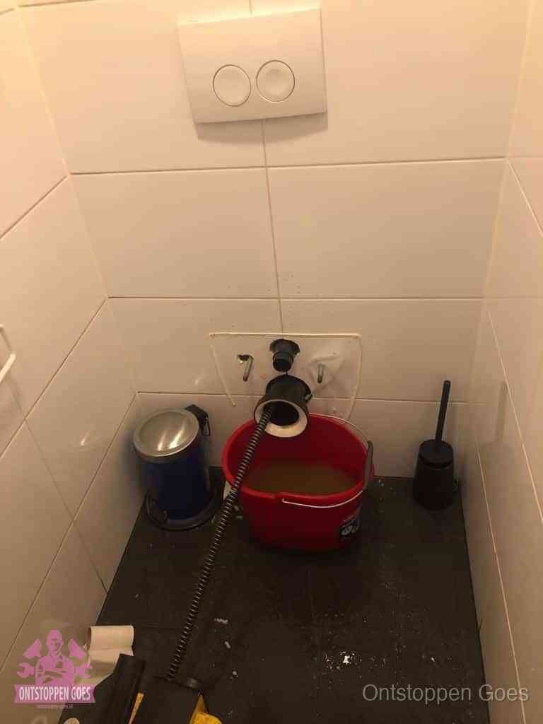 Een ontstopte wand-wc met verwijderde toiletpot waar een ontstoppingsveer in de wanduitgang verdwijnt, het achterhalen waar de verstopping zit in Goes