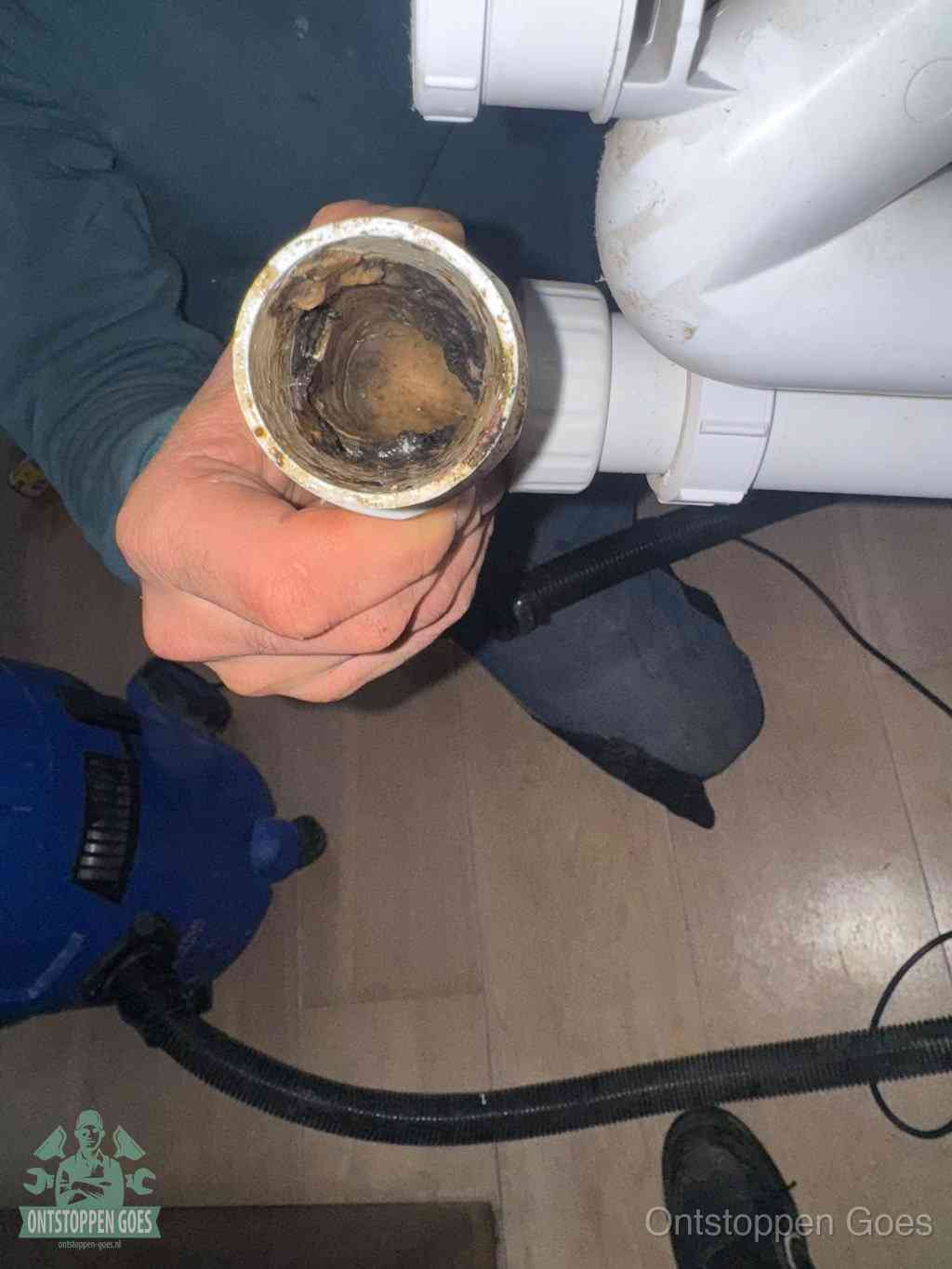 Een hand houdt een losgekoppeld PVC-afvoerbuisstuk vast dat van binnen volledig is dichtgeslibd met vet, vuil en aanslag, de meest voorkomende oorzaak van een verstopte afvoer in Goes