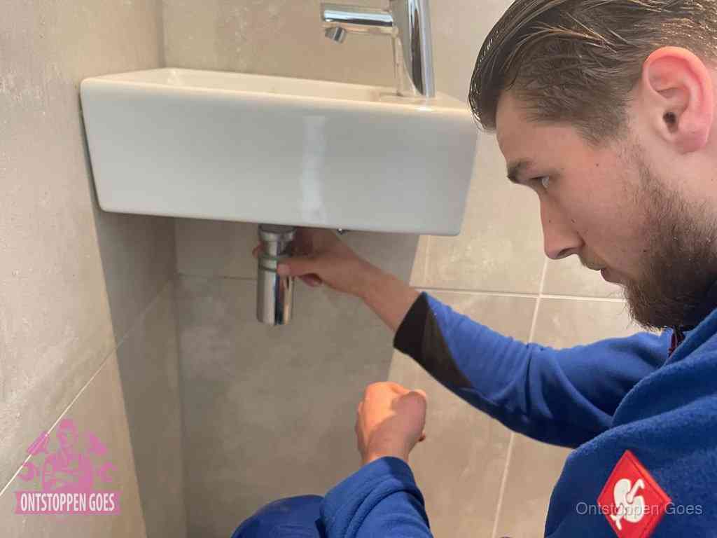 Een monteur draait met de hand de zwanenhalssifon onder een fonteinwasbak in de toiletruimte los om de bron van rioollucht in Goes te vinden